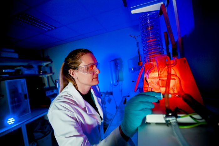 Till vänster, en kvinna i vit labbrock och skyddsglasögon. Till höger, labbutrustning av glas.