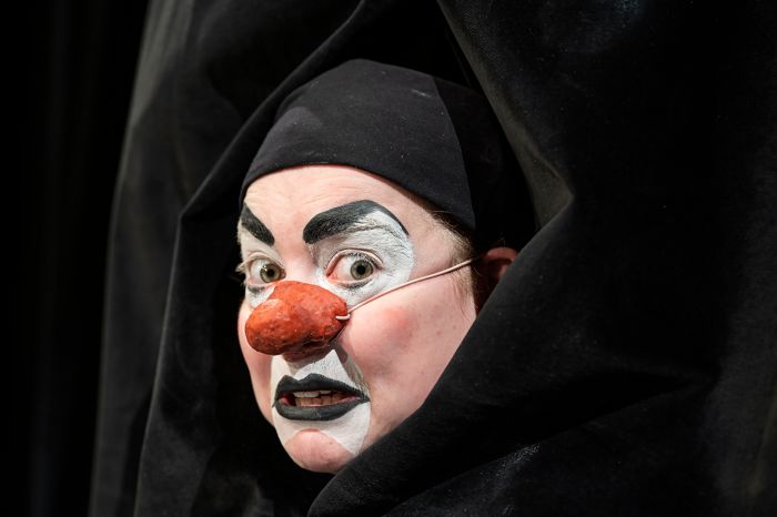 Kvinna sminkad till clown tittar ut från ett svart draperi.