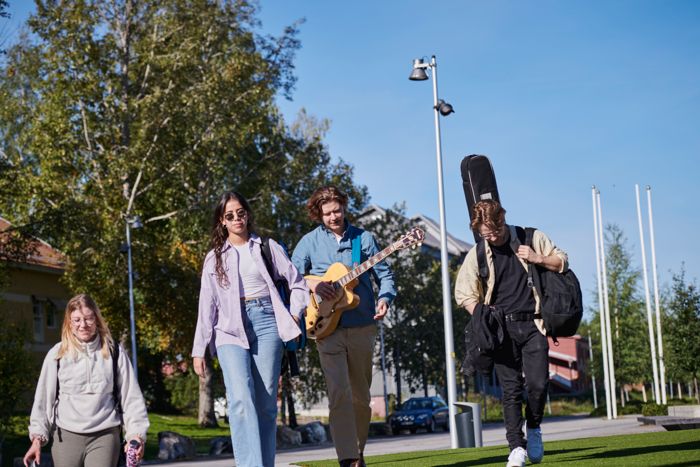 A woman and two men walking towards the camera. The man in the middle carries a guitar. The man to the right carries a guitar case.